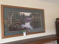 Framed signed wall art depicting a lakeside cabin scene with deer and flowers, hung on a wall above a wooden surface.
