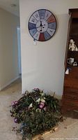 Wide angle view showing round wall clock hung on a wall above a cluster of faux plants, one real plant, and faux wreath on the floor.