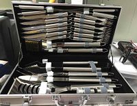 Full view of BBQ tools arranged inside carry case, showing knives, forks, skewers, brush and spatula held with elastic straps.