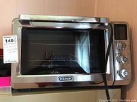 Front view of the DeLonghi electric convection oven showing stainless steel and glass door with wire rack inside.