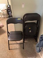 Five black metal folding chairs shown stacked and one open in front, positioned indoors on a carpeted floor next to a vacuum cleaner.