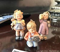 Three Goebel Hummel porcelain figures shown together, front view, located on wood surface with blurred background