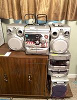 JVC CA-MX-K30 stereo system with two speakers, remote control, and antenna, shown on a wood cabinet under curtains.
