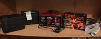 Wide shot showing contents on shelf: two black Koss speakers, red and black Century battery charger, boxed MotoMaster Eliminator battery charger with manual, decorative cover roll, and caulking spreader.
