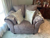 Front view of brown microsuede armchair with two throw cushions and patterned arm covers placed on armrests.