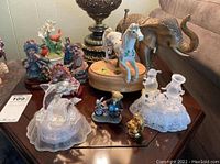 Photo of two ceramic music boxes including a carousel horse on wooden base, composite elephant, multiple figurines including Louis Dionne and Cherished Teddies, gold painted Buddha with peeling paint, and plastic lighted dolphin and snowmen figurines on table.