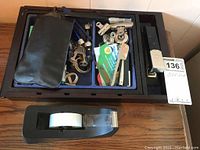 Two black trays containing assorted office accessories and one large black tape dispenser with tape roll on a wooden surface