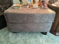 Front view of gray microsuede rectangular ottoman with dark wood feet on carpeted floor, with decorative items on top.