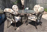 Full view of patio table surrounded by four cushioned chairs on outdoor stone patio with fence background.