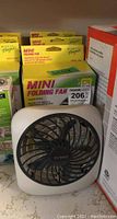 Photo showing one white square O2 COOL portable battery-operated fan with black blades, outside box, positioned in front of three unopened boxed fans.