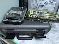 Three cases shown stacked. Black plastic 50 piece socket set case with transparent lid. Silver metal 40 piece ratchet socket set case open displaying sockets and tools inside. Gray plastic carrying case for Rotozip Spiral Saw at bottom.