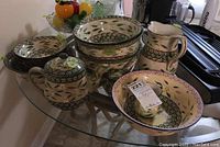 Photo shows a variety of Temp-tations ovenware pieces arrayed on a glass table, including bowls, a jug, a teapot, cream and sugar bowls, all with floral and leaf patterns in cream, green, and purple. Some chips noted on items.