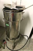 Photo of stainless steel turkey fryer pot stacked on matching lid on top of stainless steel stand with attached gas hose and regulator.