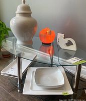 Glass table with lot items: white ceramic lidded urn on top left, Himalayan salt lamp fire bowl glowing orange, white heart-shaped napkin holder, rectangular and square white ceramic platters below, white bowl on platter.