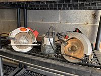 Two circular saws and one submersible pump arranged on a metal shelf with concrete block wall background, showing overall lot contents.