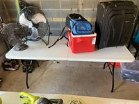 White plastic folding table with black metal folding legs in a garage setting, various items placed on top and underneath the table