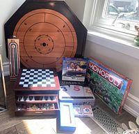 Photo showing all games and boards including crokinole board, Monopoly, chess/checkers set with drawers, puzzle, cribbage board, Rummikub, card decks, and dominoes