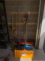 View of gardening tools including orange and yellow snow shovels, pruning shears, rake, and other hand tools against a concrete block wall.