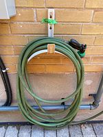Front view of coiled green garden hose weathered condition on white hanger mounted to brick wall with sprayer nozzle attached.