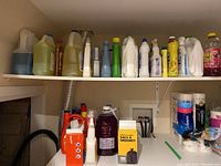 Shelf with assorted bottles of laundry detergent and cleaning supplies, including a large purple liquid jug, yellow detergent bottle, spray bottles, and boxed garbage bags.