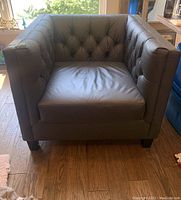 Front view of gray leather armchair showing tufted back and sides, single seat cushion, standing on wooden floor near window.