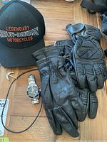 Top-down photo showing Harley-Davidson black cap, pair of men's medium black leather gloves, pair of women's large black leather biker gloves, Invicta wristwatch, and anchor necklace with leather cord.