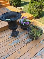 Outdoor shot of the cast iron urn and two black plastic planters, one with blooming flowers and the other with dry plants, placed on peeling painted wooden deck.