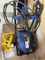 Front view of blue Simoniz pressure washer showing engine cover label and hose wrapped on frame