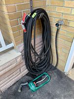 Photo showing the coiled 50-foot black rubber hose attached to an outdoor faucet, two spray nozzles including a red one attached to hose and one gray nozzle on the ground, and a green Gilmour oscillating sprinkler on pavement beneath hose.