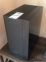 Black Samsung monitor speaker placed on a dark wood surface featuring a fabric front grille and top control buttons.