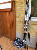 Van Bergen skis with poles leaning against brick wall; rollerblades and ski boots at base