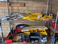 Image shows metal shelving with extension cords on top shelf, Motomaster battery jumper cables, yellow electrical connector organizer box, flashlights, and red toolbox with hand tools below.