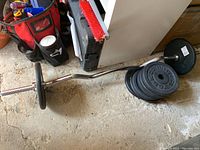 Weights and barbell set lying on a concrete floor, showing six black weight plates stacked on one side of the bar and two stacked on the other.