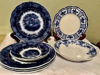 Top view of all eight flow blue dishes including plates, saucers, scalloped plate and shallow bowl arranged to show pattern and sizes.
