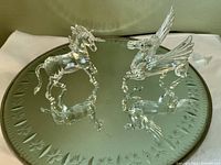 Two clear crystal figurines, a Pegasus with wings and a standing unicorn, displayed on a round mirror etched with a floral pattern.