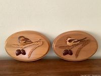 Two oval intarsia wood plaques with chickadees and acorn leaf branches mounted on wall.