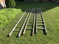 Three aluminum ladder sections laid side by side on green grass, showing full length and condition, ends with orange caps.