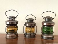 Photo shows set of 3 vintage nautical metal lanterns with colored glass (2 amber, 1 green) side by side on a wooden surface.