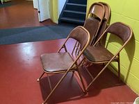 Four brown vintage metal folding chairs stacked and folded against a yellow wall on a red floor surface with carpet nearby.