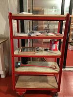 Full view of the red custom made heavy duty steel 5-tier utility cart holding a shovel, paint can, framed pictures, and small tools and hardware.