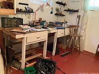 Wide view of workspace showing wooden workbenches with drawers, mounted tool racks, red adjustable desk lamp, chair, and window providing natural light.