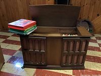 Front view of vintage Philco Ford stereo console credenza cabinet with carved wooden panels, open top lid showing inside compartments and assortment of knobs and switches, vinyl albums stacked on top.