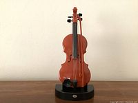 Front view of miniature violin mounted on black base, showing strings, bow, tuning pegs and metallic plate on base.