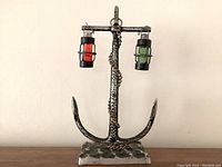 Front view of the nautical anchor shaped metal cruet set, showing the base, vertical shaft with rounded arms holding two glass bottles, one red and one green, hanging by metal cages and chain