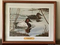 Framed artwork showing two ducks swimming on still water with surrounding aquatic vegetation. Visible gold plaque with artist name and title. Full frame and mat shown.