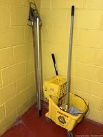 Photo showing the yellow mop bucket with wringer, two metal handles with mop cages, and mop with handle against a yellow wall.