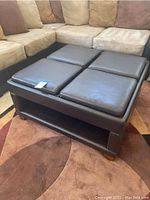 Square brown leather ottoman shown from angle, with four removable cushioned trays on top, revealing storage underneath. The cushions and leather surface show wear and creasing.