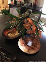 Zoomed out photo showing the orange orchid floral centerpiece in an ornate vase and the two decorative bowls, one marbled orange and beige, the other a swirled glass bowl with gold and brown tones.
