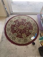 Round floor rug with burgundy center and beige floral border laid on carpeted floor.
