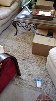 Photo showing a beige shag area carpet underneath a glass top metal frame coffee table, flanked by beige sofas and brown furniture, with cardboard boxes partially on the rug.